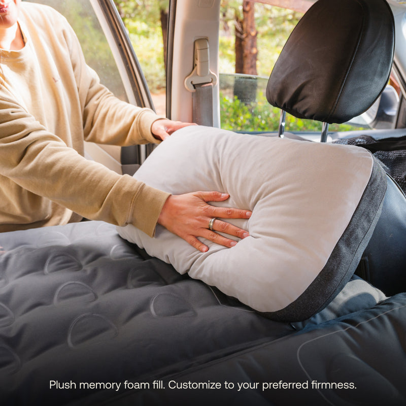 Someone adjusts a Luno HQ Packable Camp Pillow (Pre-Owned) on the backseat of a car. The seat is draped with a gray mattress pad as sunlight spills through the windows. Text reads: "Plush memory foam fill. Customize to your preferred firmness." Enjoy portability with USA shipping available.