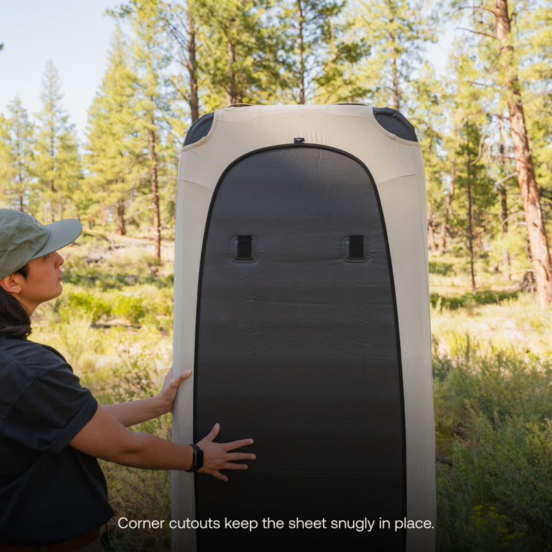 A person in outdoor gear is standing beside a tall, rectangular gray object with corner cutouts. They’re in a forest setting with trees and greenery in the background. Text at the bottom reads, "Corner cutouts keep the breathable Luno® Fitted Mattress Sheet - AIR+FOAM Pro Camping Mattress snugly in place.