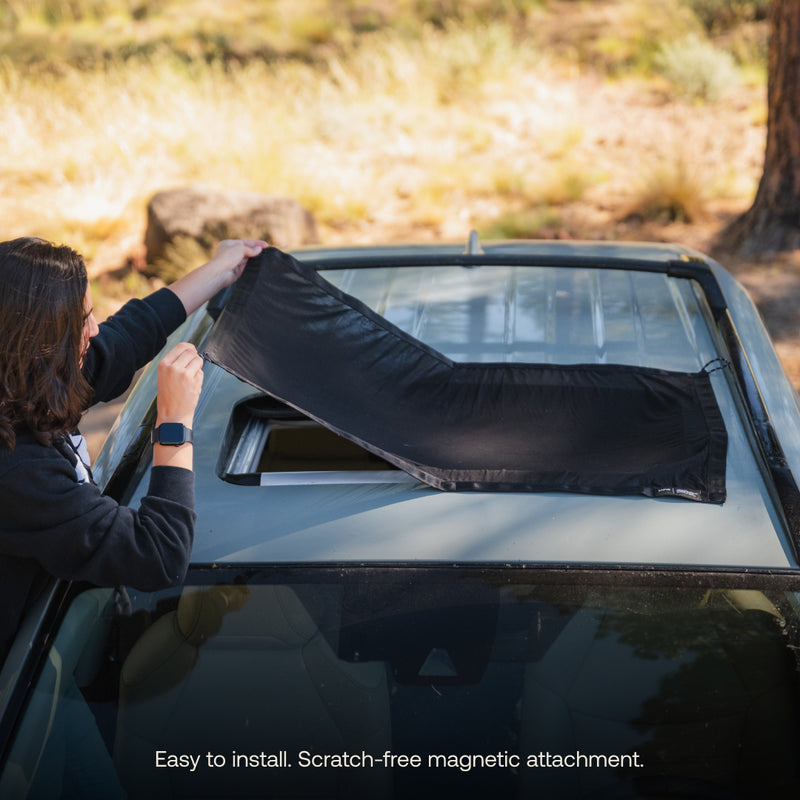 A person with long hair is setting up a black Luno HQ Sunroof Window Screen (Pre-Owned) outdoors. The text states, "Easy to install. Scratch-free magnetic attachment. Block insects while maintaining airflow." Trees and grass can be seen in the background.