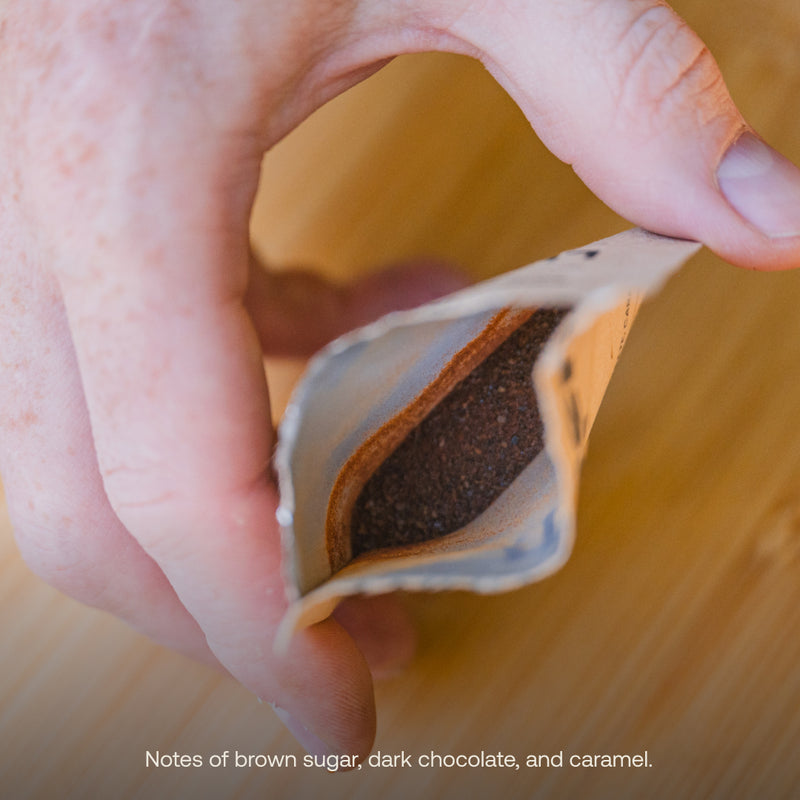 A hand is holding an open packet of ground coffee on a wooden surface. The text at the bottom reads, "Notes of brown sugar, dark chocolate, and caramel." Discover the perfect travel-friendly coffee for outdoor adventures – Campers Blend Instant Craft Coffee by Luno®.