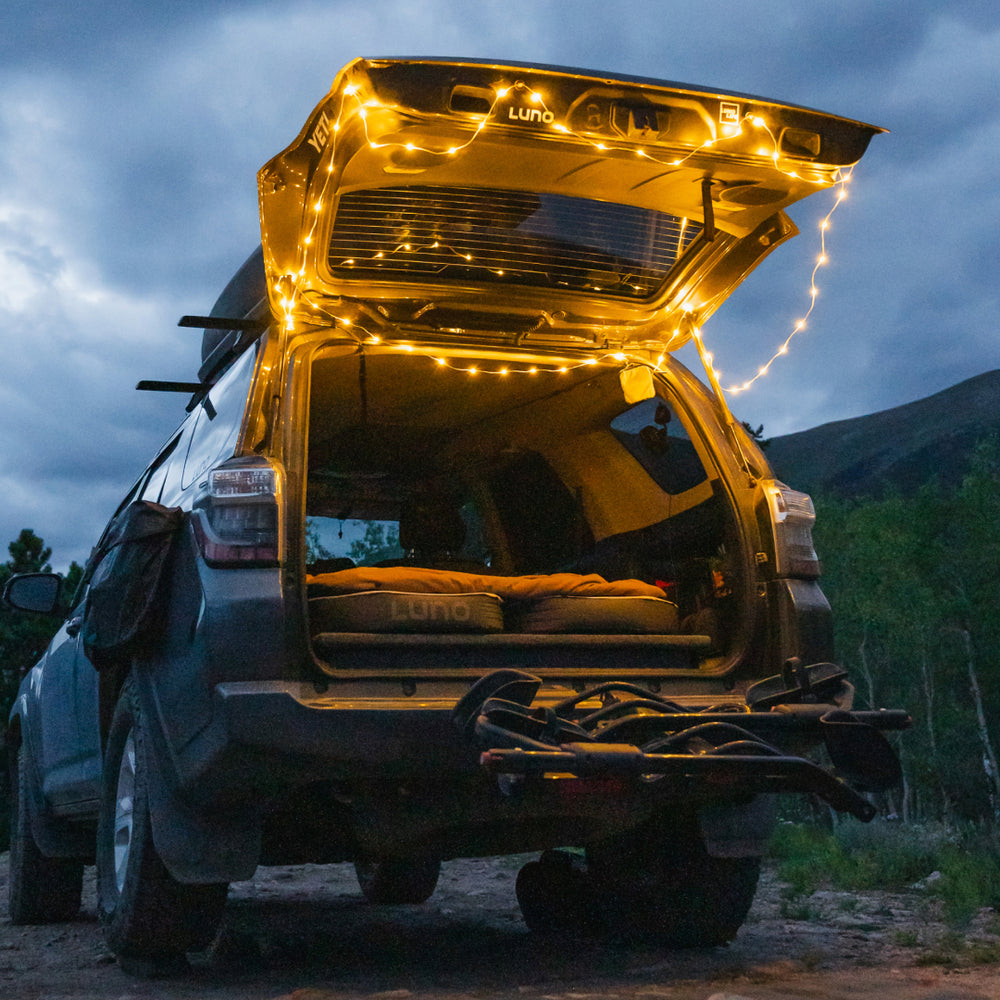 A parked SUV with its tailgate open, revealing a cozy setup with a mattress inside. Luno® Camping String Lights are draped around the tailgate, emitting a soft glow with adjustable brightness. The vehicle is surrounded by trees under a cloudy, dusk sky, powered by a rechargeable USB-C battery pack.