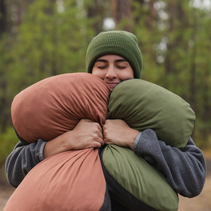 In an outdoor setting, a person wearing a green beanie and gray jacket embraces two colorful Luno® Camping Pillowcases made from cooling jersey knit. With eyes closed and an expression of contentment, the serene vibe is enhanced by the blurred view of trees in the background.