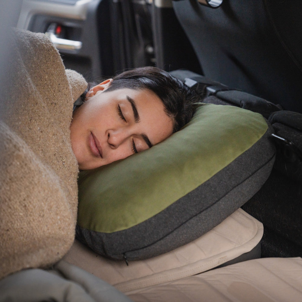 A person sleeps peacefully in a car, resting their head on a Luno® Camping Pillowcase made of cooling jersey knit. A beige blanket drapes over them, and the cozy interior of the vehicle is partially visible.