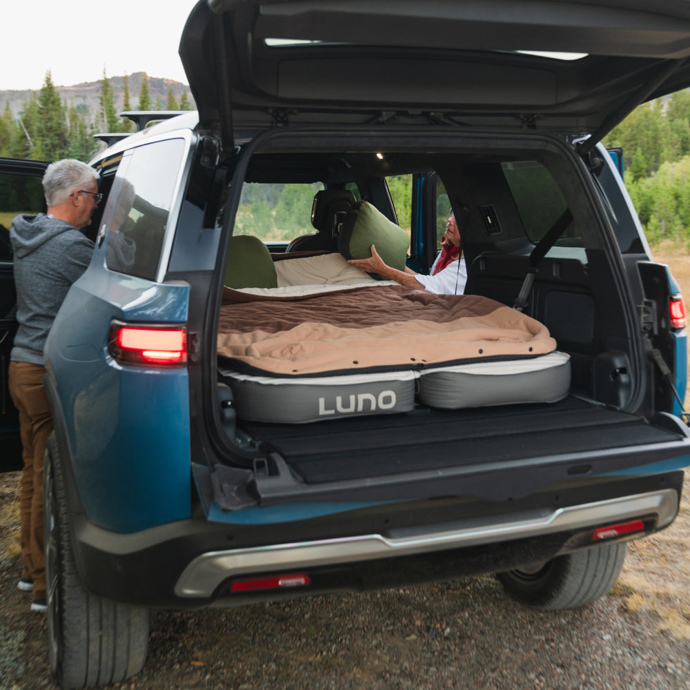 Two people set up an AIR+FOAM PRO Rivian R1S Mattress by Luno® in the back of an SUV, parked outdoors with trees and hills in the background. The vehicle's trunk is open, showcasing Luno's innovative sleep system inside.