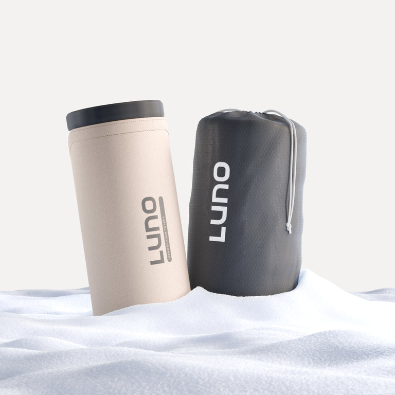 On a snowy surface, two items from the Luno® Car Camping AIR Comfy Bundle make their presence known: a tall, beige cylindrical mug with a black lid and a black drawstring bag, both adorned with the white Luno logos. They are perfect companions for setting up your car camping mattress in nature's icy embrace.