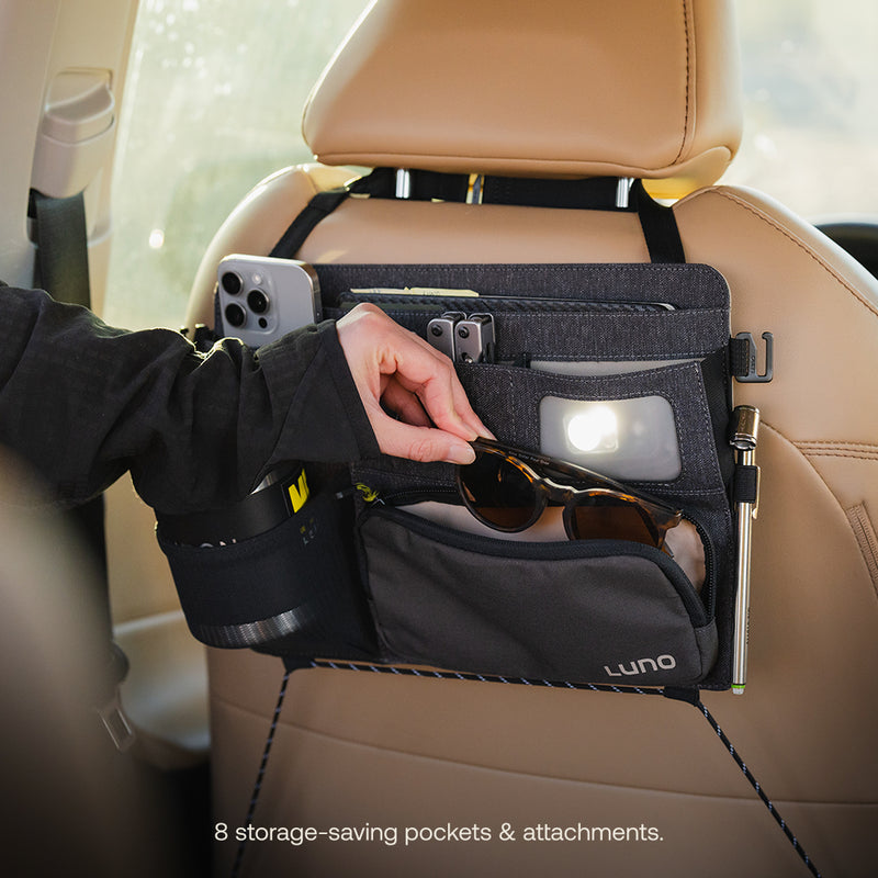 A person stores sunglasses in a Seatback Organizer by Luno®, attached to a tan car seat. Ideal for vehicle organization, it features 8 storage-saving pockets and attachments to hold a phone, flashlight, pen, and mug.