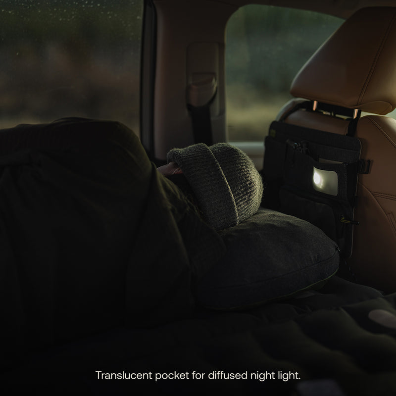 A person in a beanie sleeps in a car's back seat at night. A dim light from the Luno HQ Seatback Organizer (Pre-Owned) softly illuminates with its translucent pocket, perfect for road trips. Available for USA shipping.