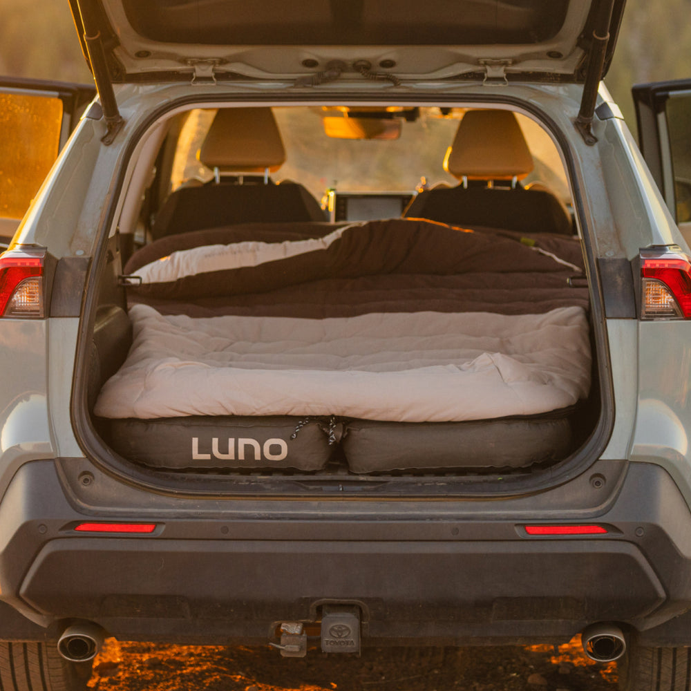 The car trunk becomes a cozy sleeping area with a Luno® Fitted Sleeping Bag Comforter and vehicle mattress, ideal for car camping. The rear hatch opens to reveal a scenic outdoor sunset backdrop.