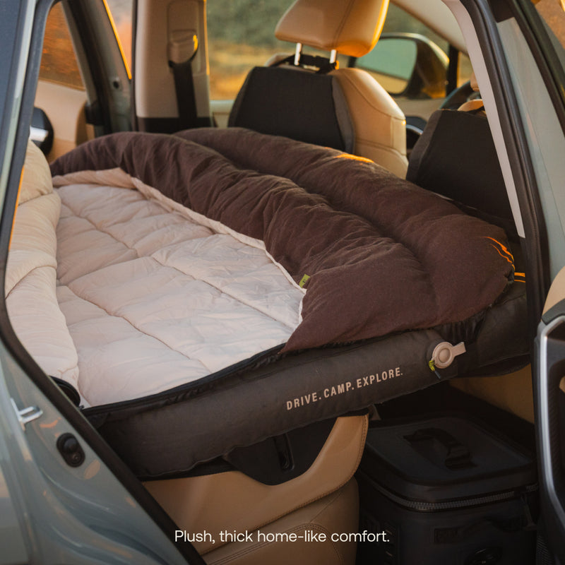A car is set for camping with a Luno® Fitted Sleeping Bag Comforter, offering cozy, home-like comfort across the backseat. Text on the comforter reads "DRIVE. CAMP. EXPLORE.