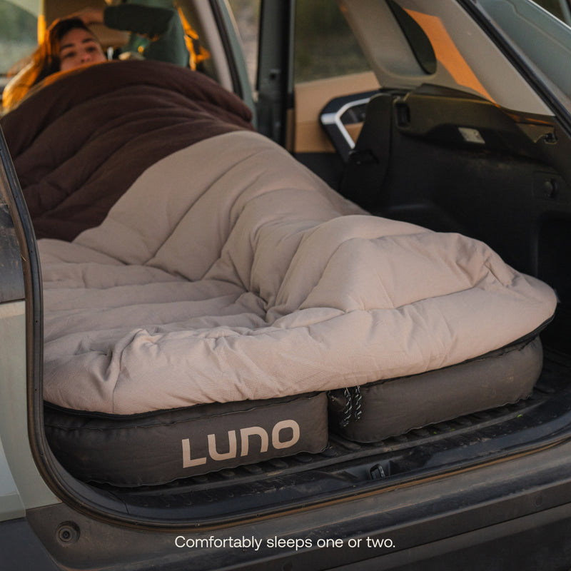 A person is lying under a Luno® Fitted Sleeping Bag Comforter on an inflatable mattress inside a car's rear compartment. The snug fit creates a cozy sleeping area, and sunlight filters in, adding warmth.