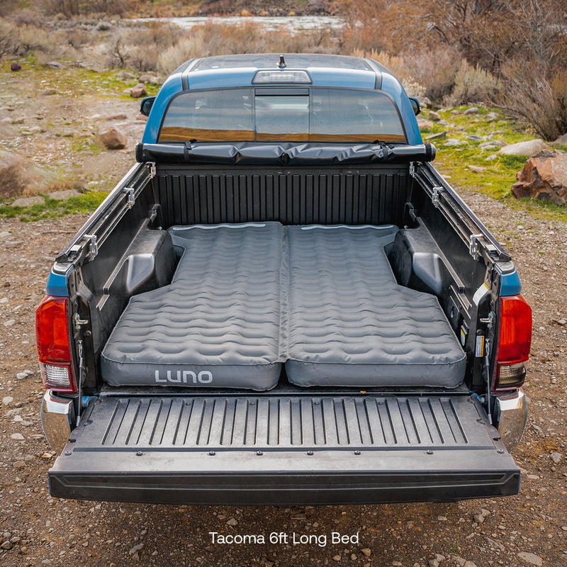 A blue pickup truck with its tailgate down showcases an inflated, pre-owned "Luno HQ AIR Truck Bed Mattress" snugly fitted inside its 6-foot bed. Parked on a dirt path amid rocky terrain and dry bushes, it embodies a resourceful spirit in its rugged, repurposed setup.