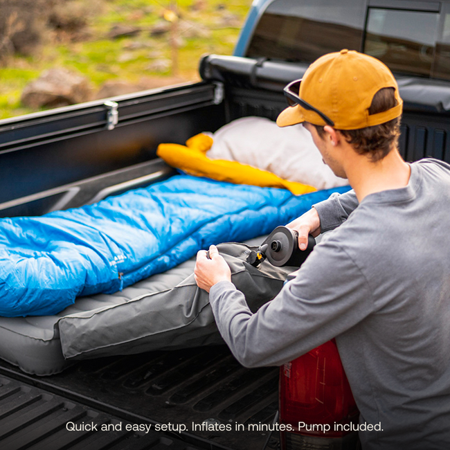 Someone wearing a cap is inflating an AIR Truck Bed Mattress by Luno HQ in the back of a pickup truck. Nearby, pre-owned gear such as a blue sleeping bag and pillow are arranged on it. The truck is parked outdoors on rocky terrain, ready for adventure.