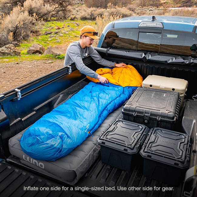 A person sets up the blue AIR Truck Bed Mattress (Pre-Owned) from Luno HQ in a truck bed, wearing a hat and sunglasses. The truck holds black and white storage cases. Caption: "Inflate one side for a single-sized bed; use the other side for gear." Enjoy USA shipping!.
