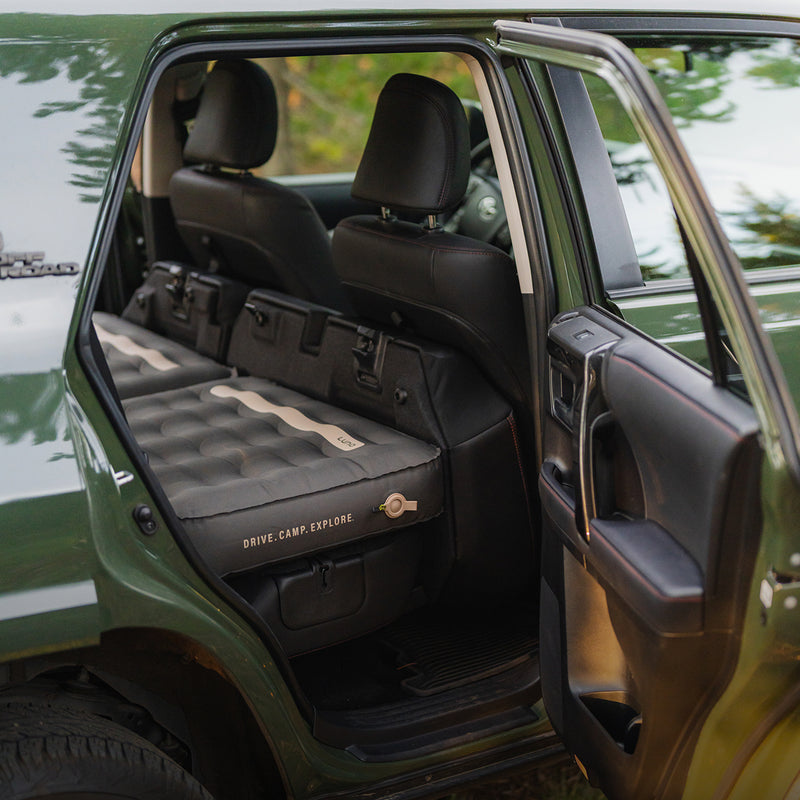 A Luno® AIR Pro 4Runner Vehicle Mattress is set up in the backseat of a green SUV, with the rear door open and front seats pushed forward, ready for car camping adventures.