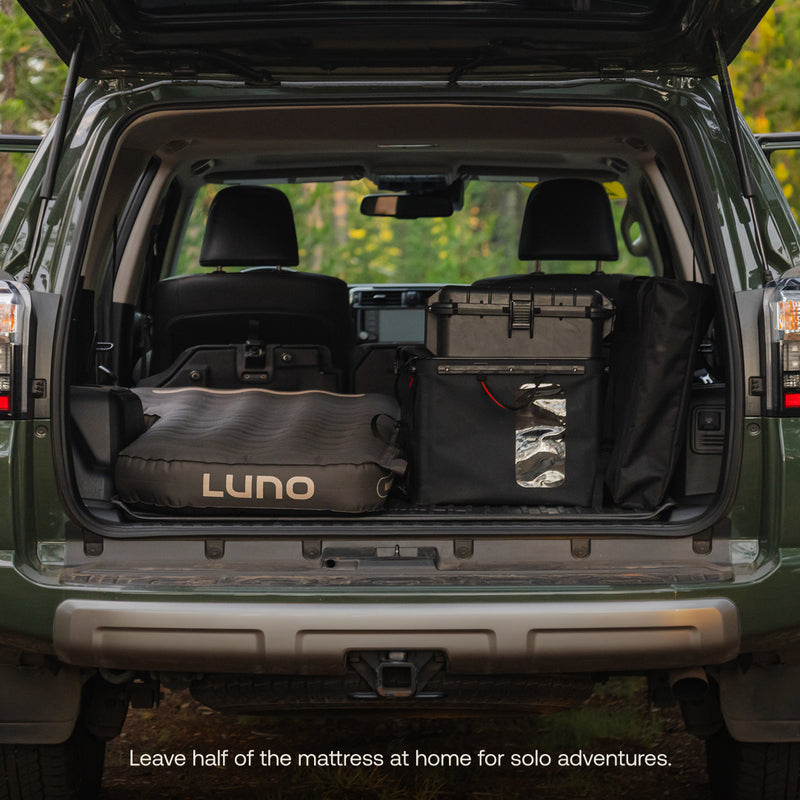 With the rear seats folded, a Luno® AIR Pro 4Runner Vehicle Mattress fits on one side of the SUV trunk, leaving space for gear on the other. Trees are seen through the windshield. Text: “Leave half of the mattress at home for solo adventures.”.