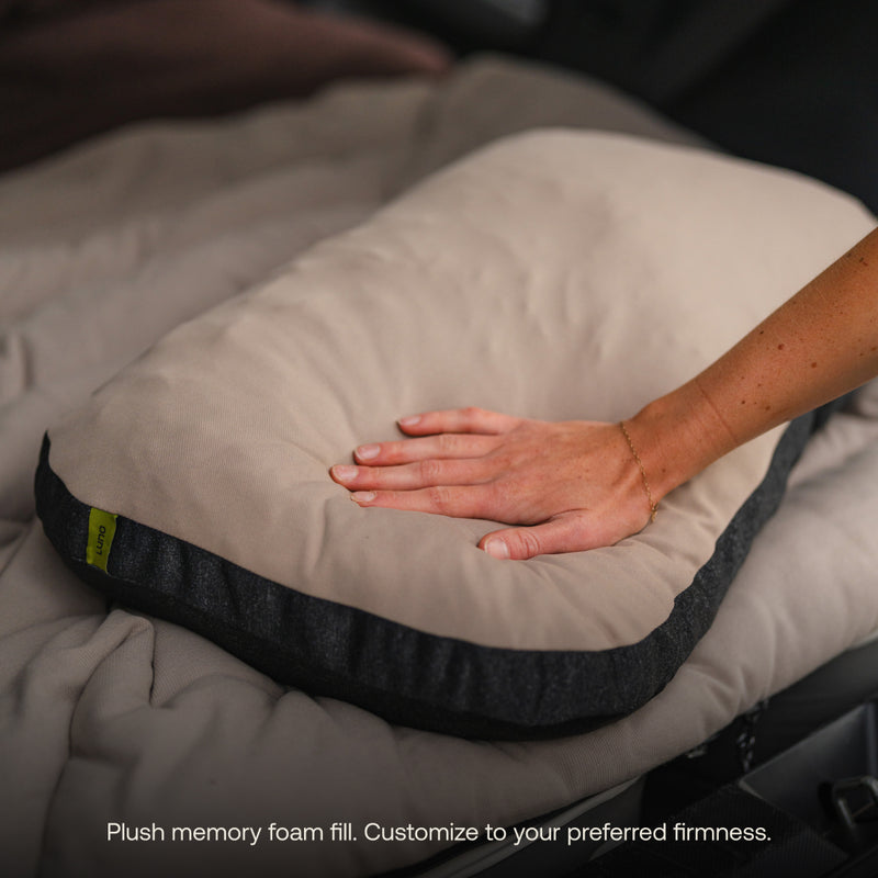 A hand presses on the Luno® Packable Camping Pillow in beige and dark gray, highlighting its softness. The pillow sits on a quilted surface. Text below: "Plush memory foam fill. Customize firmness to your preference.