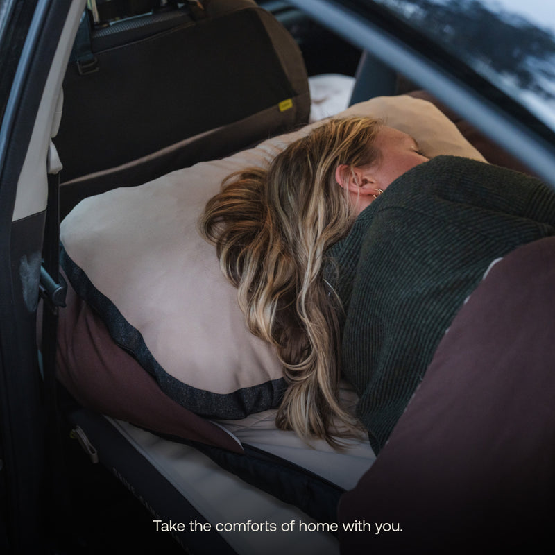A person with long blonde hair relaxes on pillows and blankets in a car’s backseat. Caption: Bring the comfort of home anywhere—explore the AIR BASE Car Camping Sleep Kit for your next adventure.