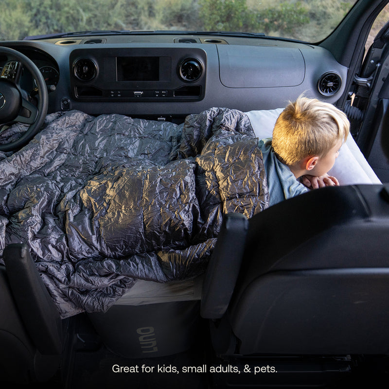 A young child relaxes under a gray blanket on an AIR Pro Van Front Cab Mattress (Pre-Owned), set up across the vehicle's front seats, gazing out the passenger window with the dashboard and steering wheel in view.