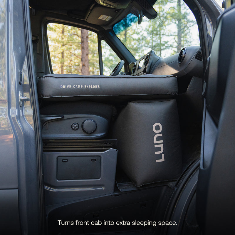 A van interior featuring the Luno® AIR Pro Van Front Cab Mattress, set up above the front seats to create a comfortable cab bed; text at the bottom reads, "Turns front cab into extra sleeping space.