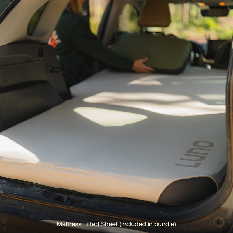A person places a green pillow on an inflated AIR BASE Car Camping Sleep Kit with a fitted sheet inside the back of an SUV—perfect for vehicle camping. Text below reads: Fitted Sheet included in bundle.