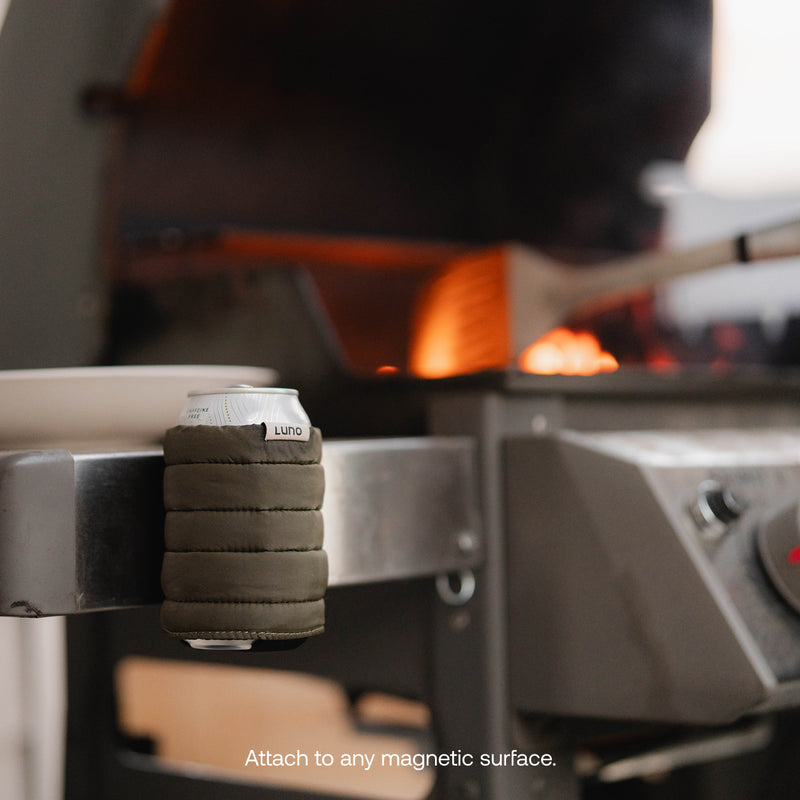 A Magnetic Can Cooler securely holds a soda can to the metal side of a grill, with grill flames flickering behind and a spatula resting nearby.