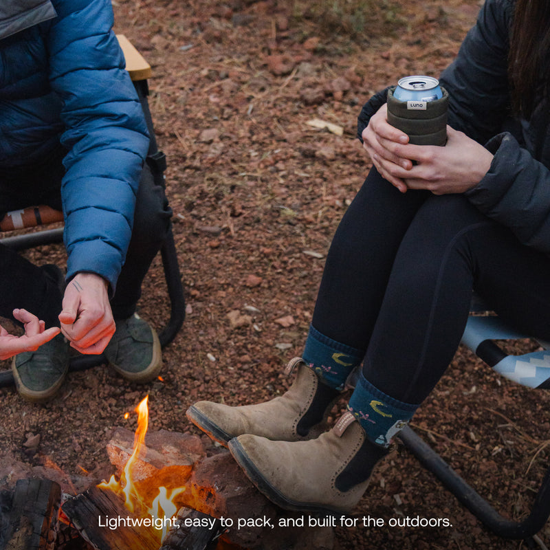 Two people in outdoor gear sit by a campfire on rocky ground. One holds a mug, the other gestures; a Magnetic Can Cooler rests nearby. Text reads: Magnetic Can Cooler—lightweight, easy to pack, and built for the outdoors.
