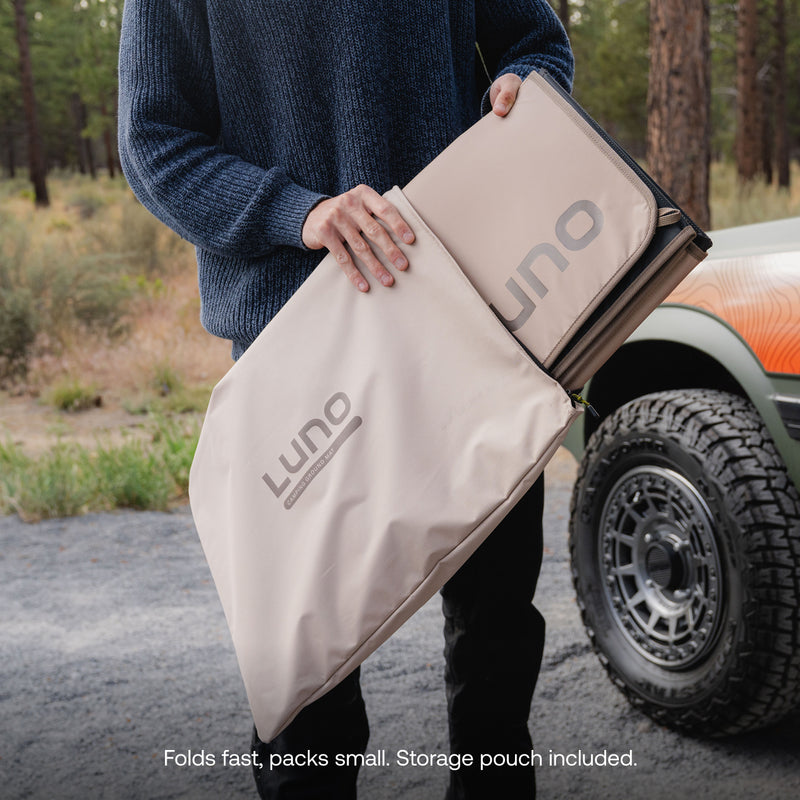 A person holds a folded beige Camp Ground Mat (3x5’) with its storage pouch outdoors near a car, ready for adventure. Perfect as a car camping mat or portable outdoor mat, it folds quickly and packs compactly—storage pouch included.