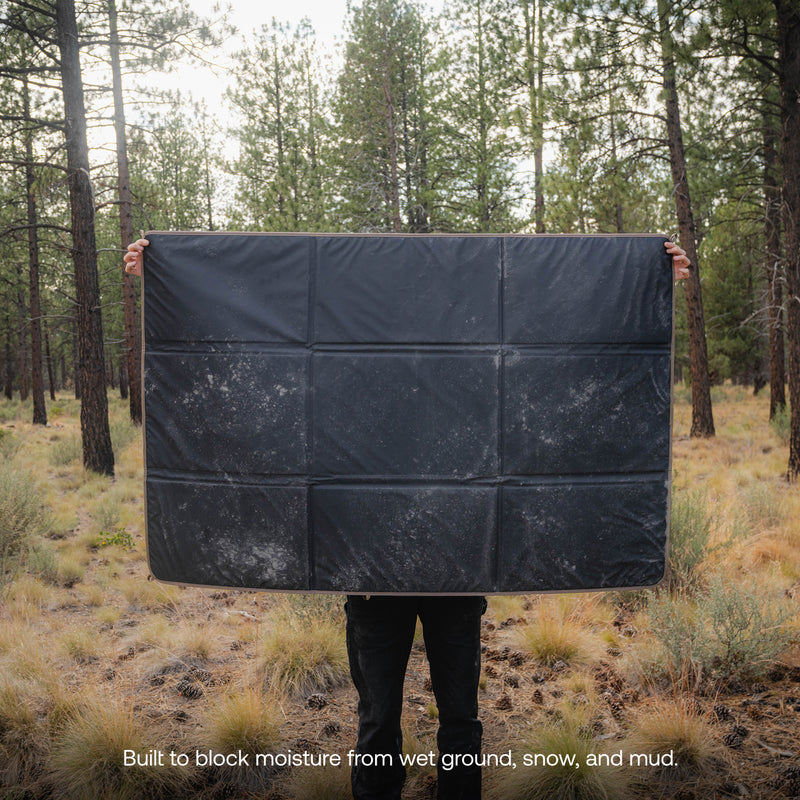 A person stands in a forest holding the Camp Ground Mat (3x5’), a large black waterproof mat with visible dirt marks that covers most of their body. A caption reads: Built to block moisture from wet ground, snow, and mud.