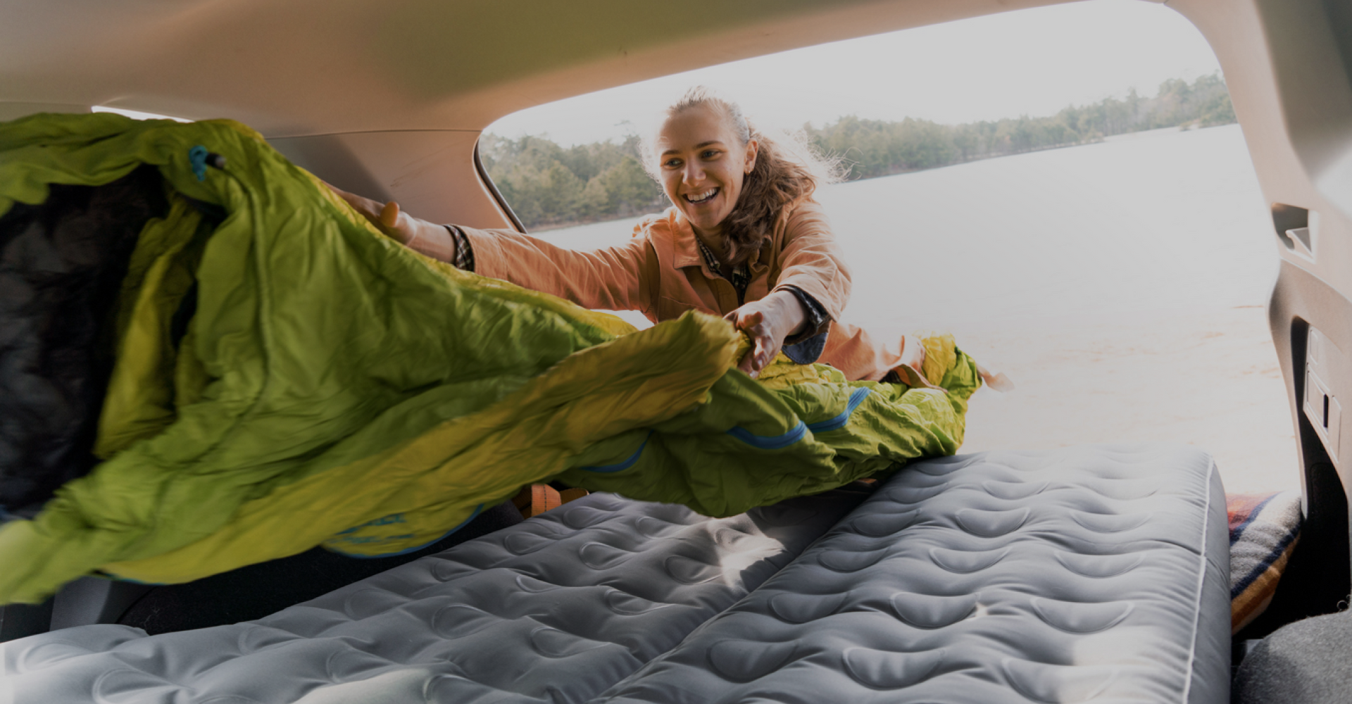 A person smiling while laying out a green sleeping bag onto an inflated mattress in the back of a vehicle. Trees and a body of water are visible in the background outside the vehicle.