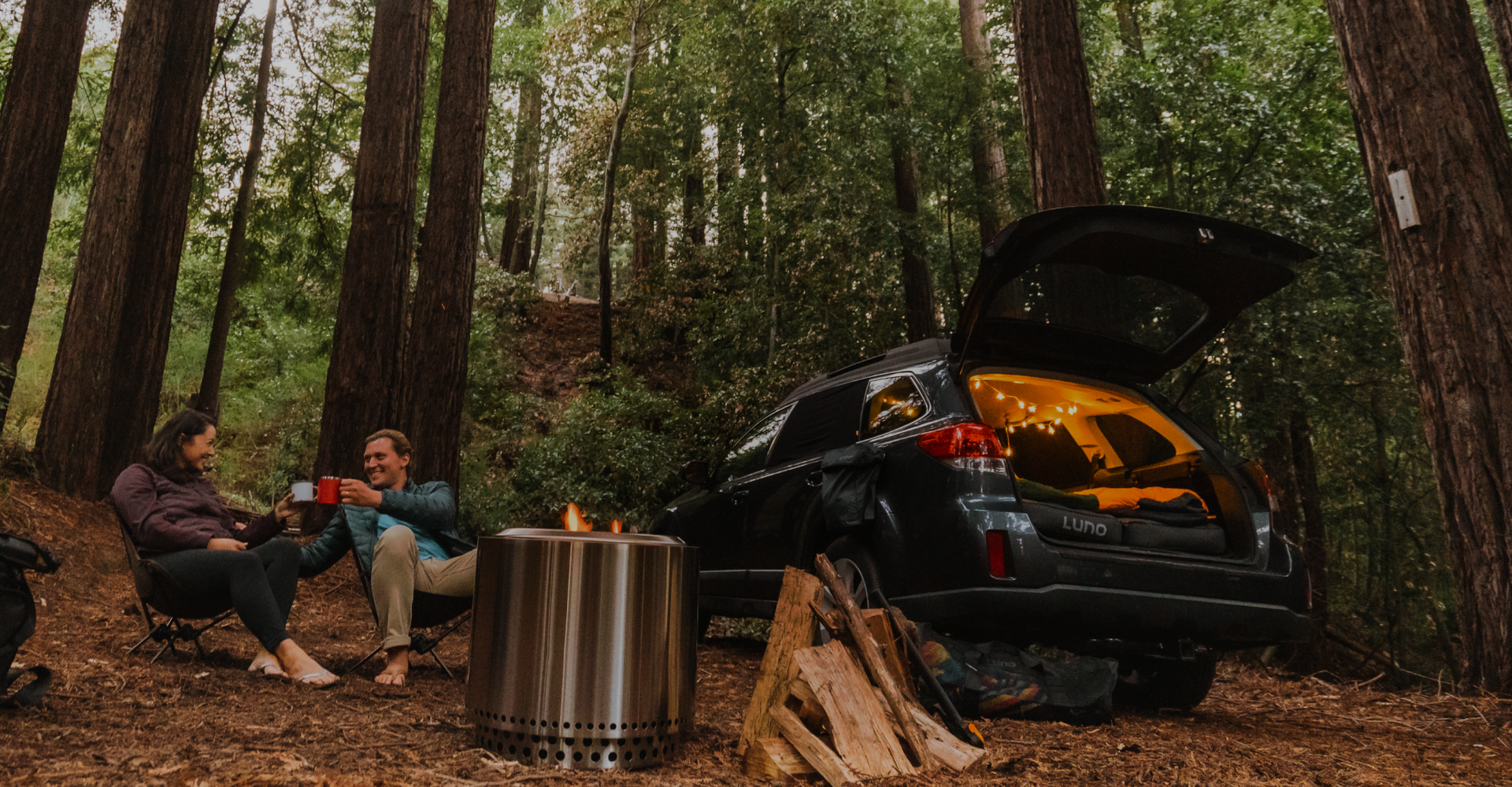 A man and woman sit on camp chairs in a forest, smiling and holding mugs. Behind them, a car with an open hatchback reveals its lit interior. A portable fire pit burns beside them, with stacked logs nearby. Tall trees surround the cozy campsite.