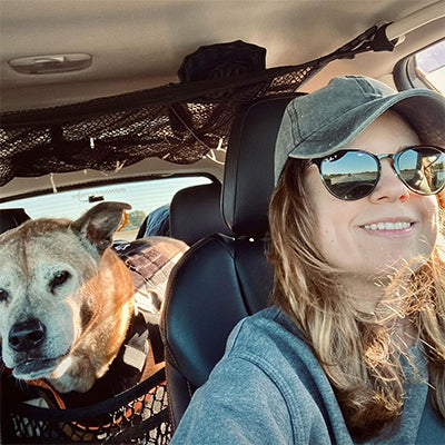 A woman wearing sunglasses and a baseball cap is smiling while driving a car. In the back seat, a dog with a light-colored coat is looking forward. A net holds items overhead in the car. Sunlight is streaming in through the windows.