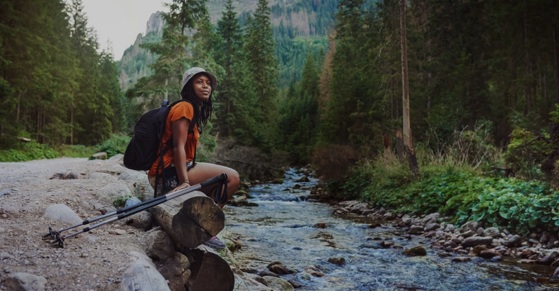 A hiker wearing an orange shirt, black shorts, and a hat sits on a log next to a river in a forested mountain area. They have a black backpack and walking sticks and look off into the distance. The scene is lush with green trees, rocks, and flowing water.