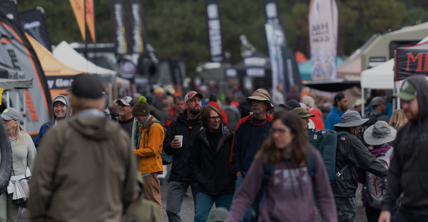 A crowded outdoor gear festival or market with many people wearing jackets, hats, and backpacks. Various vendor tents and banners line both sides of the path, displaying outdoor-related items and activities under an overcast sky.