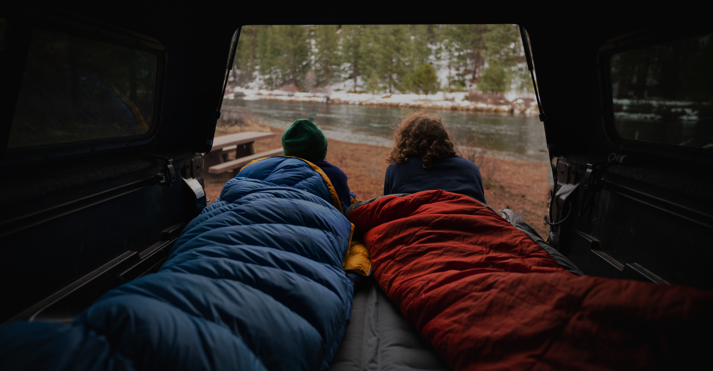 Two people are lying in sleeping bags, one blue and one red, in the back of a vehicle with an open hatch, overlooking a tranquil forest river. Snow patches are visible on the ground and trees, suggesting a chilly outdoor setting. Both are gazing out peacefully.