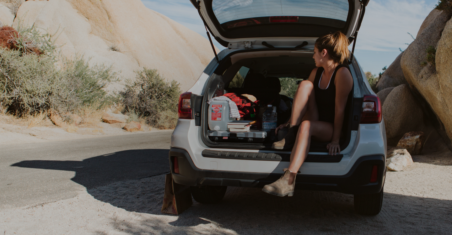 A person sits in the back of an open SUV in a rocky desert area. Camping gear and a cooler are in the trunk. The person, wearing a tank top and shorts, has a relaxed posture and looks out at the landscape, which features large boulders and sparse vegetation.