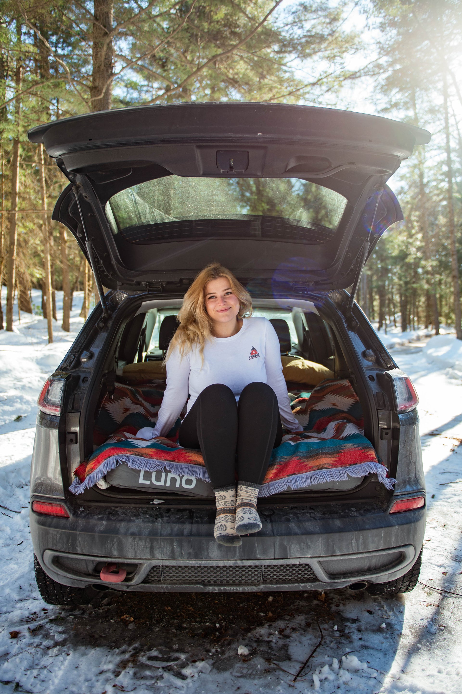 A person with long blond hair is sitting on the edge of an open car trunk in a forested, snowy area. They are wearing a white long-sleeve shirt, black pants, and patterned socks, with a colorful blanket and camping gear inside the car. Sunlight filters through the trees.