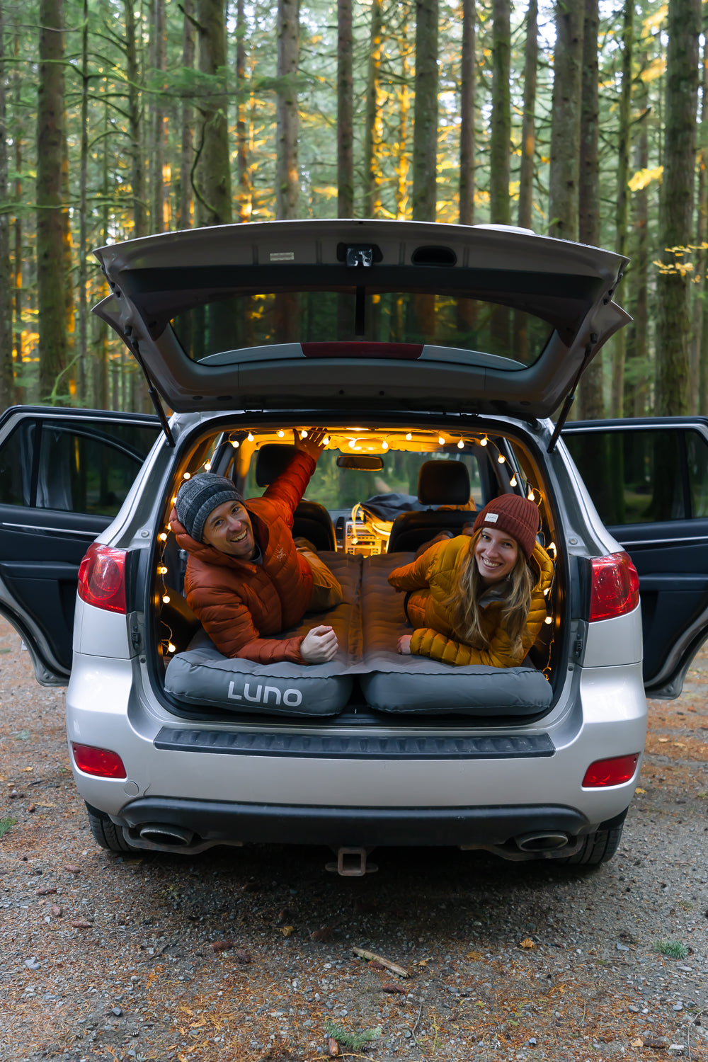 A cheerful couple relaxes in the back of an open SUV, which is parked in a forest. They are lying on an air mattress and are surrounded by string lights, creating a cozy atmosphere. Both are wearing warm clothing, and the SUV's rear seats are folded down for extra space.