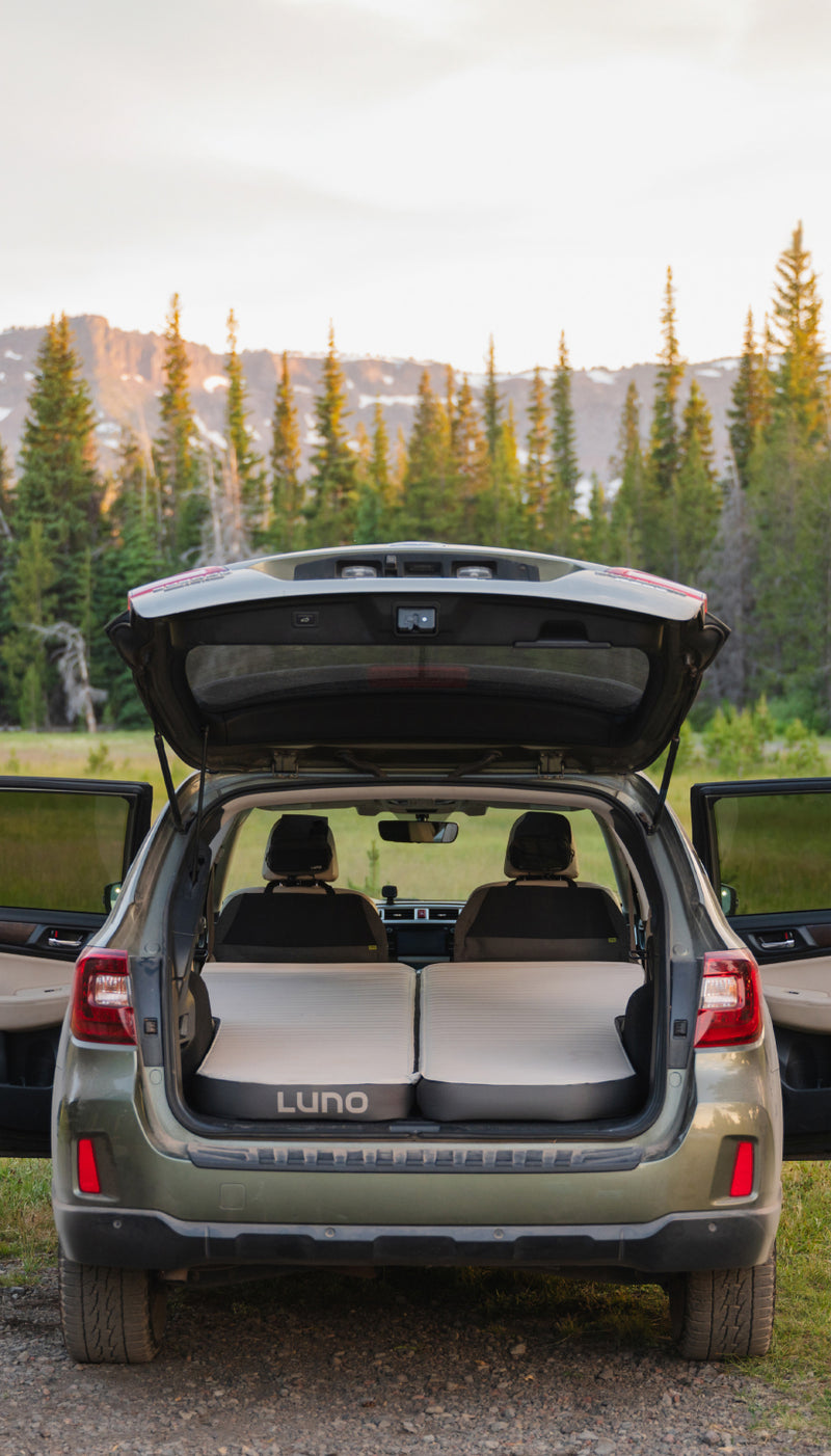 A green SUV with its rear hatch open reveals a car camping setup featuring a Luno air mattress in the back. The scene is set in a grassy area with pine trees and a mountain range in the background under a clear sky.