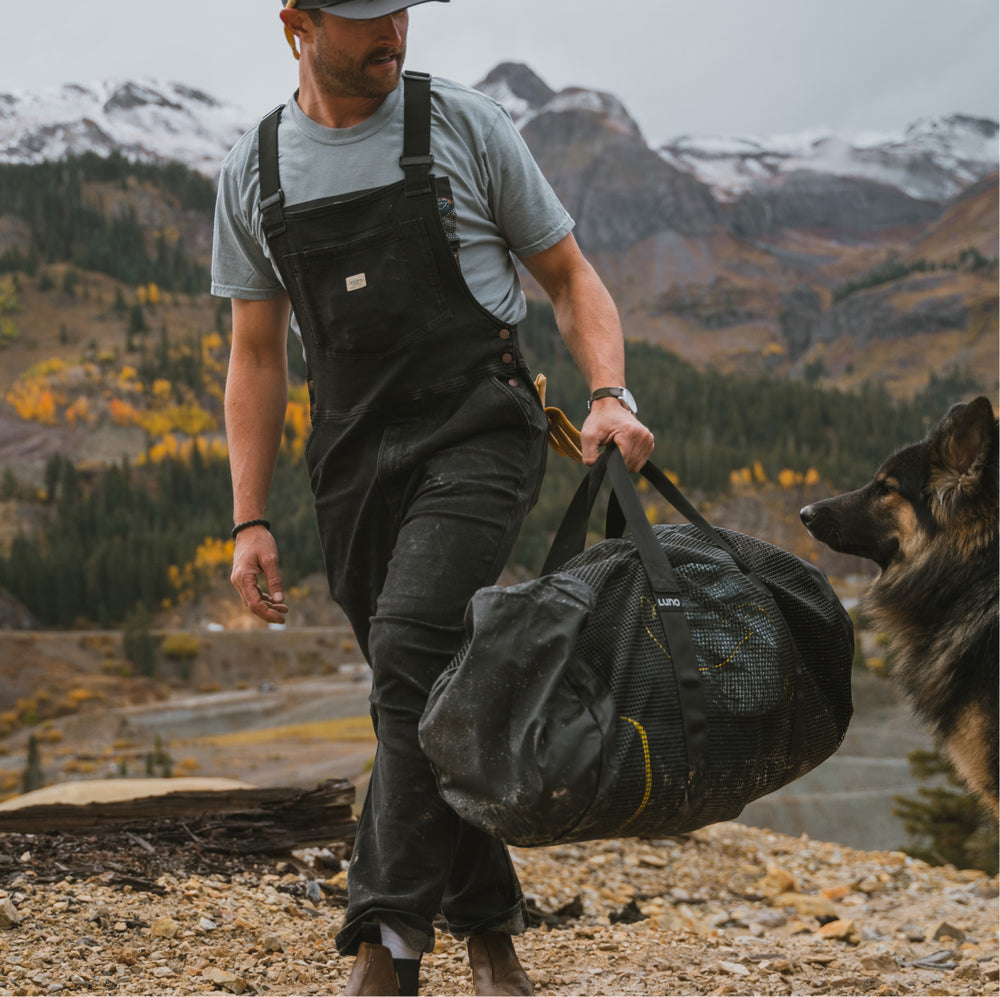 Clad in a cap and black overalls, someone carries a Luno® Mesh Gear Duffel, valued for its packable design and durable fabrics, through snow-capped mountains. A dog is nearby on rocky terrain amidst an autumnal backdrop.