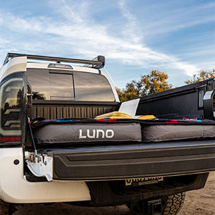 A white pickup truck with an open tailgate is parked outdoors under a blue sky. In the truck bed, an inflatable mattress branded "LUNO" is set up, covering the entire surface. Trees are visible in the background.