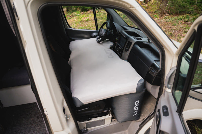 A van's interior viewed from the open driver's side door, showing an inflatable mattress covering the area from the dashboard to the front seats. The mattress is branded with "Outo" and set up for sleeping. The background shows a green forested area.