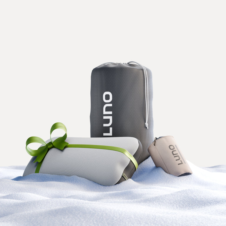 A winter-themed image features three sleeping essentials on the snow: a large gray bag, a beige cylindrical pouch, and a rectangular package wrapped with a green ribbon. Ideal for a cozy campsite, all the items are from the Camper Van AIR Sleep Bundle by Luno®.