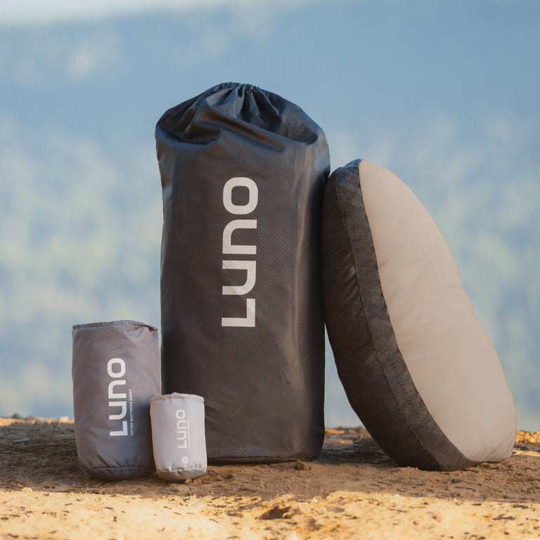 The Luno® Car Camping AIR Starter Bundle, featuring four camping gear bags and a round cushion—ideal car camping essentials—is displayed outdoors on the ground with a blurred natural landscape in the background.
