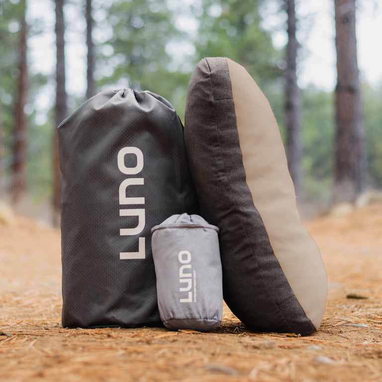Three Luno® Camper Van AIR Pro Starter Bundles—essential for van life—stand upright on a pine needle-covered forest floor, with tall trees and lush campsite greenery blurred in the background.