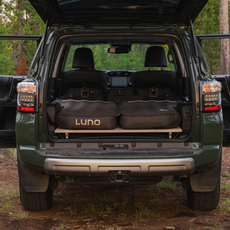 A black SUV with its rear hatch open displays a Vehicle Sleeping Platform with an inflatable mattress set up in the cargo area for car camping in a wooded outdoor setting.
