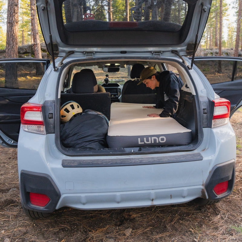 Someone is adjusting a Luno HQ AIR+FOAM PRO Camping Mattress (Pre-Owned) in the back of an SUV parked in a wooded area, highlighting its excellent condition. A sleeping bag with a helmet on top is placed next to the mattress. The SUV's rear and side doors are open, revealing tall trees in the background.