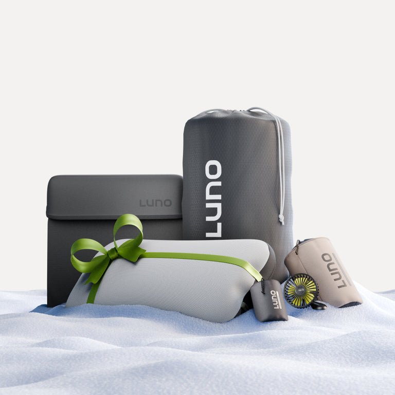 The Camper Van AIR Ultimate Bundle by Luno® is showcased on a soft surface, featuring an array of travel essentials perfect for van life. This bundle includes a stylish gray bag, a neatly packaged item with a green ribbon, a sleek cylindrical case, and a compact portable fan. Every piece in this collection comes with the assurance of our 3-year warranty to provide peace of mind.