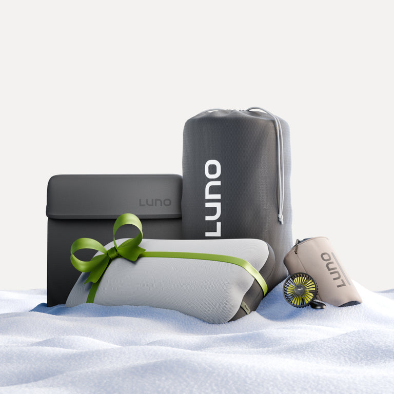 The Camper Van AIR Ultimate Bundle by Luno® features a sleek collection of travel essentials, including a gray bag, a rolled blanket with a green ribbon, and a beige container with an accompanying small brush. These items are tastefully displayed on a pristine white, snowy surface.