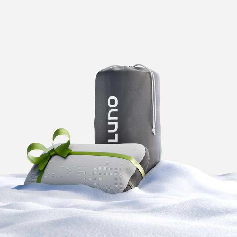 A gray bag labeled "Luno®" stands upright on a snowy surface, suggesting the Truck Camping AIR Sleep Bundle essentials. In front, a white gift wrapped with a green ribbon captures the spirit of adventure typical of winter or holiday themes.