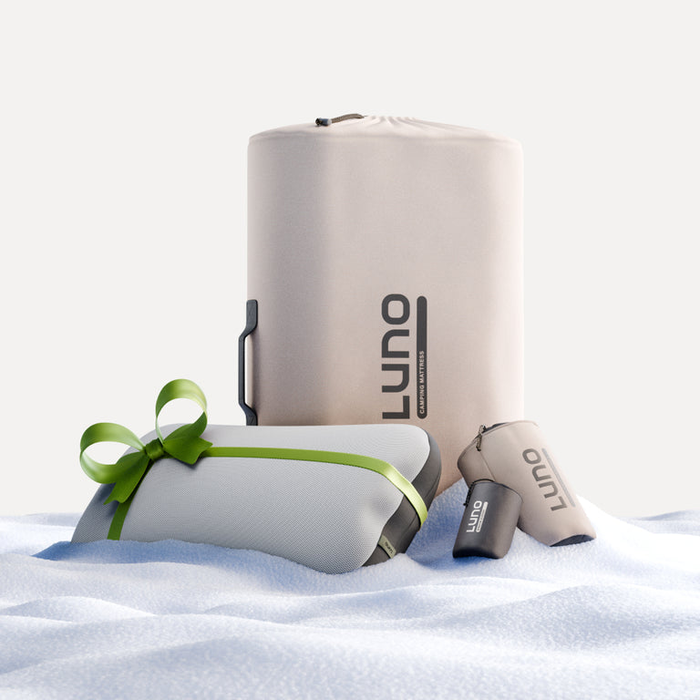 Displayed on a snow-like surface is the Car Camping AIR+FOAM Pro Starter Bundle by Luno®, ideal for car camping and comfortable outdoor adventures. This minimalist collection includes a large cylindrical bag, two smaller pouches, and a rolled-up package tied with a green ribbon, all featuring the Luno logo.